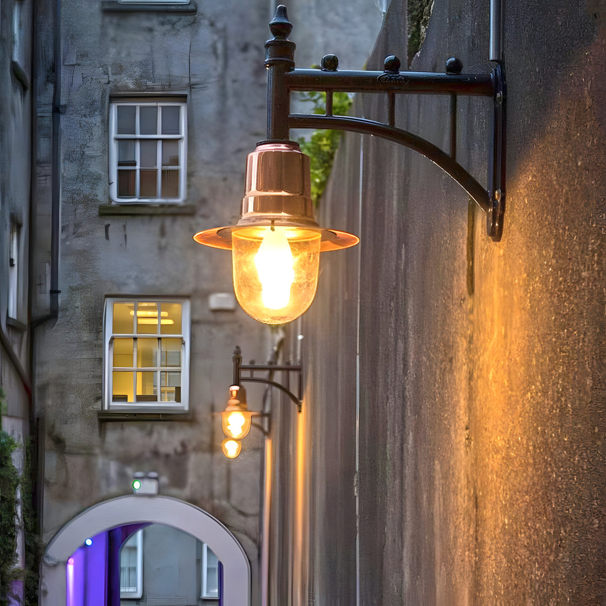 Medium Railway Style Wall Lantern on a cast arm with copper shade and clear glass dome, mounted on an exterior stone wall in a narrow alley setting.