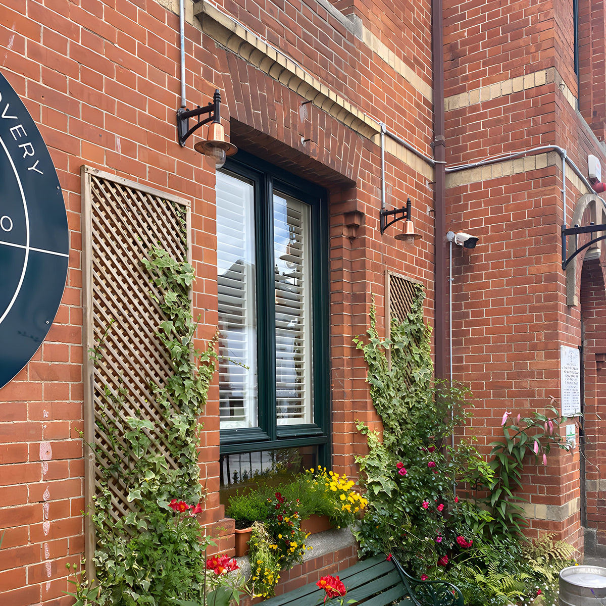 Small Railway Style Wall Lantern with copper spun shade on a black cast arm, mounted on a red brick exterior wall alongside a matching lantern and window.