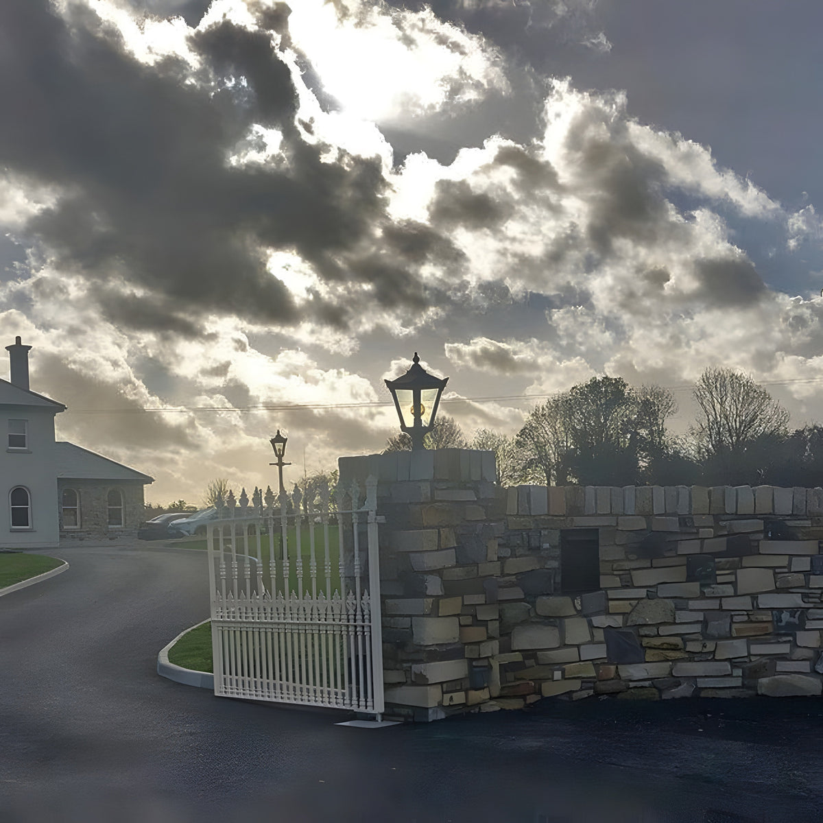 Victorian Manor cast iron pillar light in black, mounted on a stone gate pier cap with a lantern-style shade, shown at a residential entrance driveway.