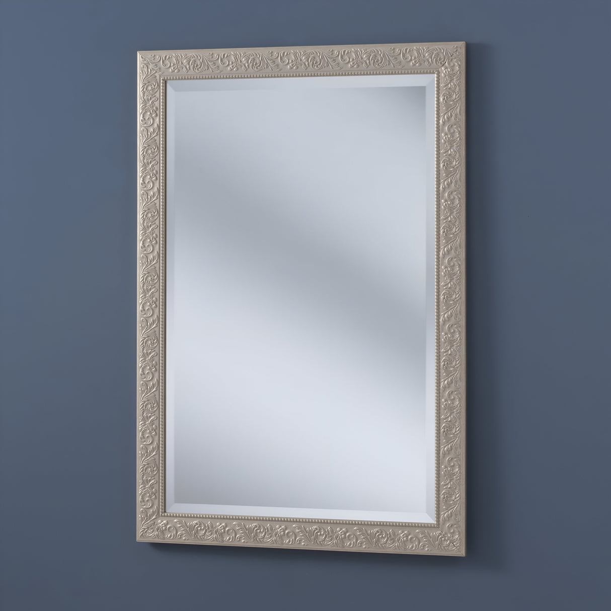 The Perleon Rectangle Wall Mirror - Silver elegantly hangs on a dark blue wall. Its ornate design acts as a decorative centerpiece, while the reflective surface softly blurs and disperses light throughout the room.