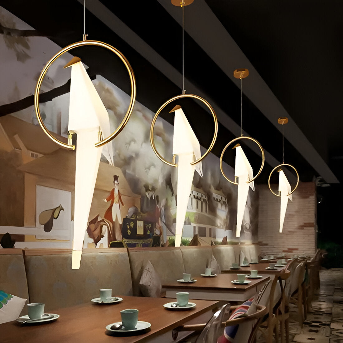 A modern restaurant interior showcases Ex-Display Bird LED Pendant Lights in White & Gold hanging stylishly above tables set with small plates and cups. An abstract mural decorates the background, enhanced by this energy-efficient lighting solution.