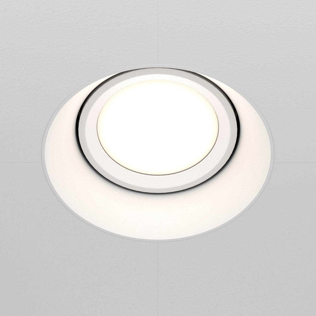 The Dot Single Tilt Plaster-in Round Recessed Downlight in white is shown in a close-up, illuminating a clean, white ceiling with its bright, even glow. Its tilt feature offers customizable lighting to complement any space.