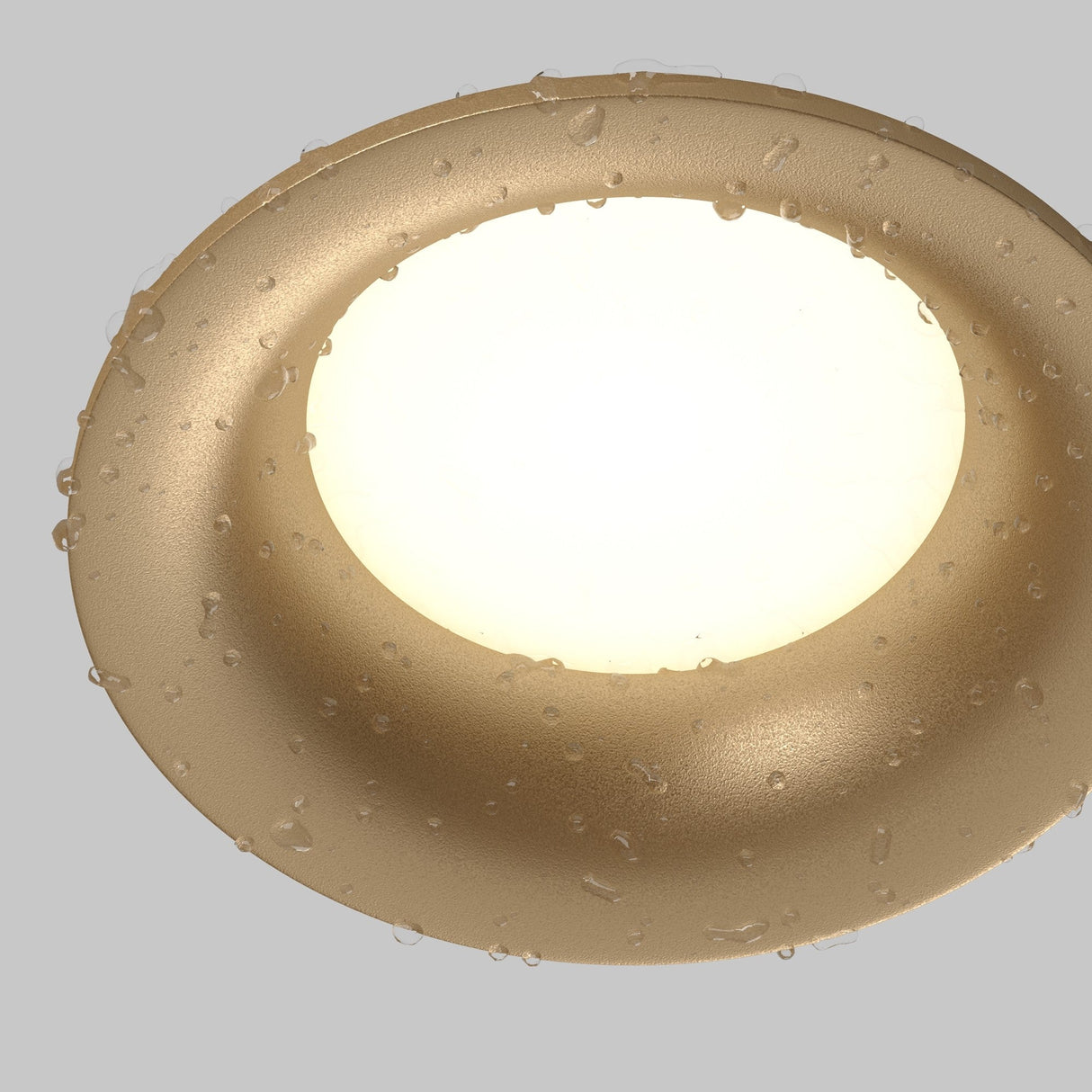 A close-up of the Slim Outdoor Fixed Round Recessed Downlight in matte gold, featuring water droplets on its surface, illuminated against a plain gray background.