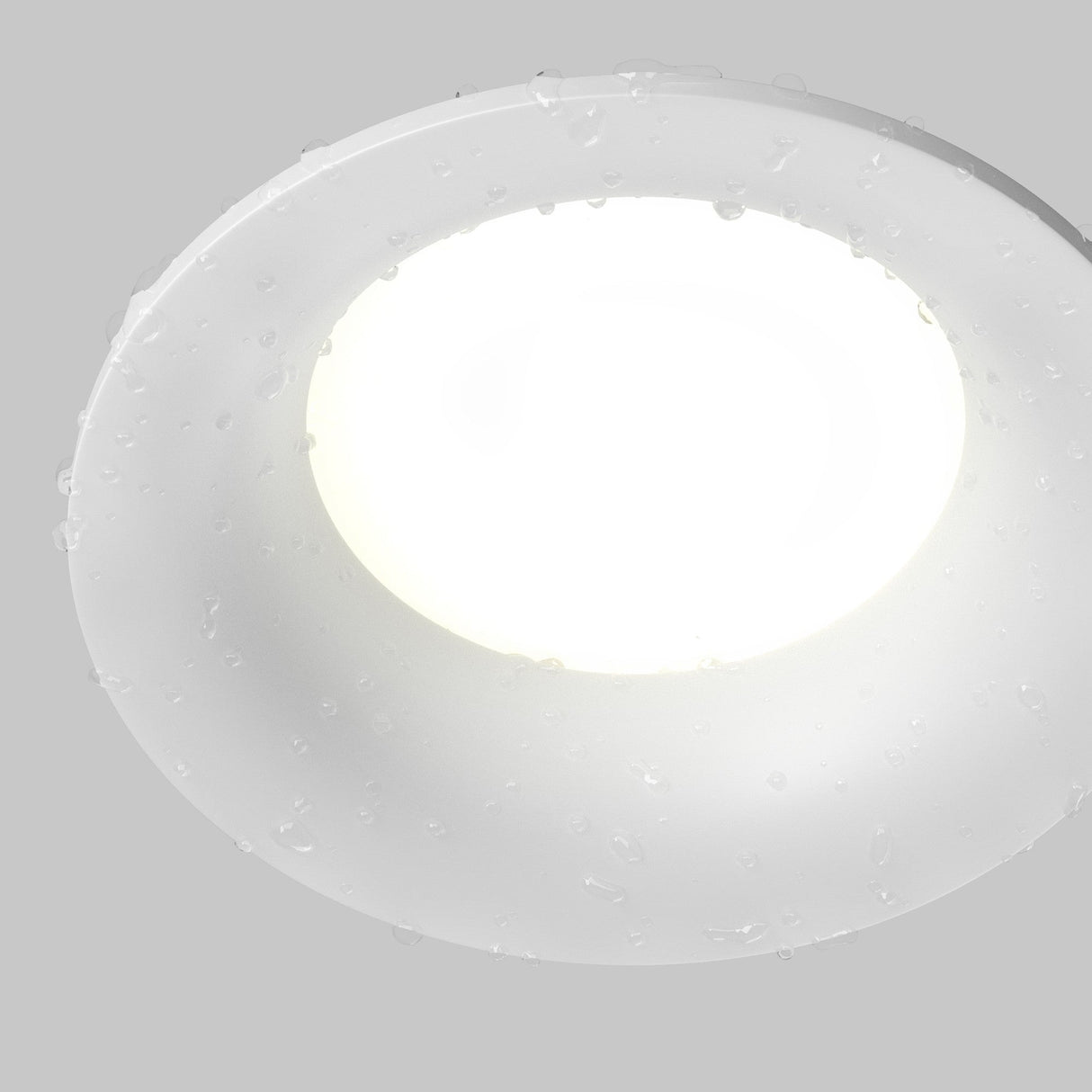 The Slim Outdoor Fixed Round Recessed Downlight in white is lit and adorned with water droplets, indicating its weather resistance, set against a plain gray backdrop.