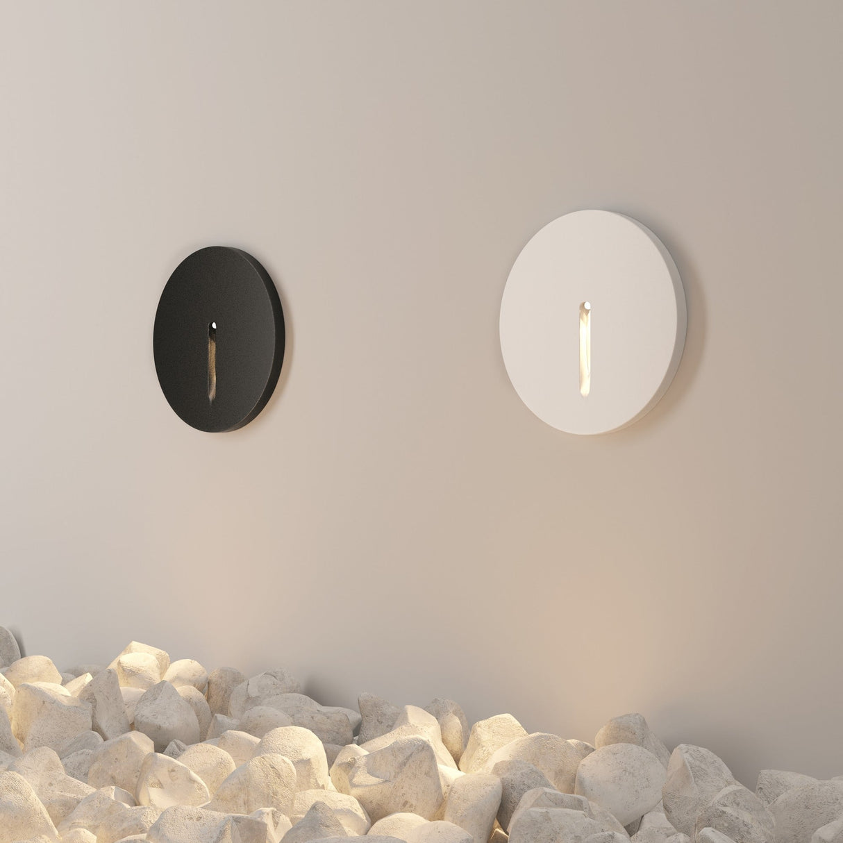 Two round wall lights, one black and one white, are mounted on a light-colored wall. Smooth white stones below are illuminated by ambient lighting from Lock Outdoor LED Recessed Wall Light - Black, creating a minimalist and serene atmosphere with durable materials for lasting elegance.