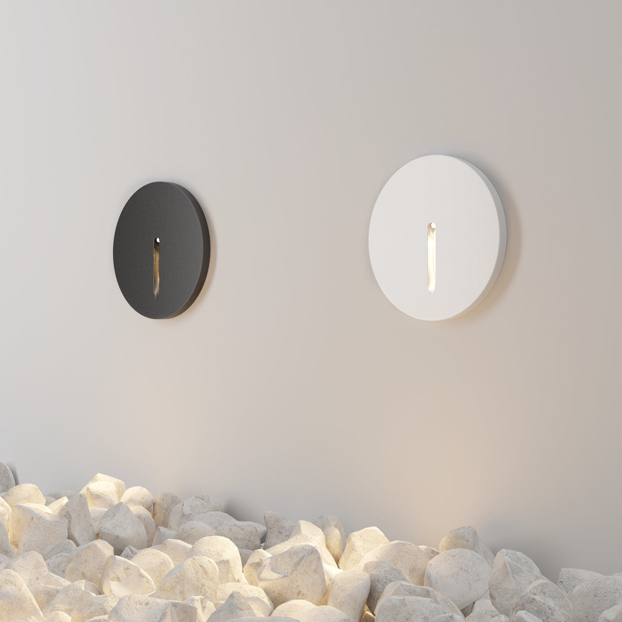 A light-colored wall above white stones features two Lock Outdoor LED Recessed Wall Lights, one black and the other white, their narrow slits emitting a soft, warm glow and creating an elegant, modern ambiance.