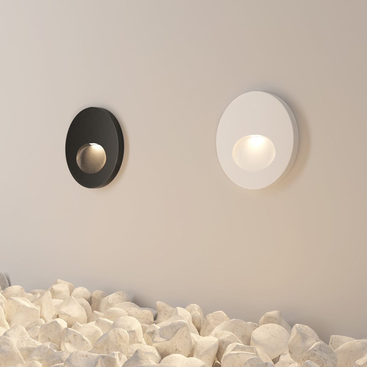 Two circular wall lamps, one black and sleek and the other white, light up a beige wall. They resemble the Bil Outdoor LED Recessed Wall Light with an IP54 rating and are set above white stones, adding to the minimalist modern decor.