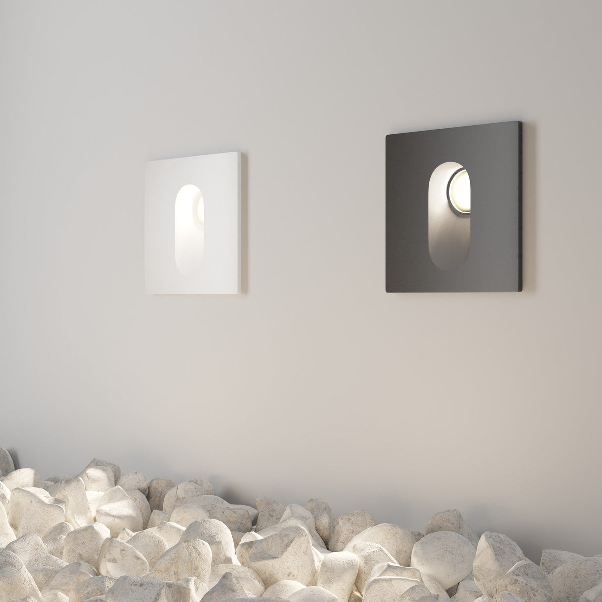 Two modern square wall lights, one white and one black, featuring the Via Urbana Outdoor LED Recessed Wall Lights IP54 rating, illuminate a wall. Below them is a bed of white pebbles enhancing the minimalist and contemporary design with its corrosion-resistant construction.