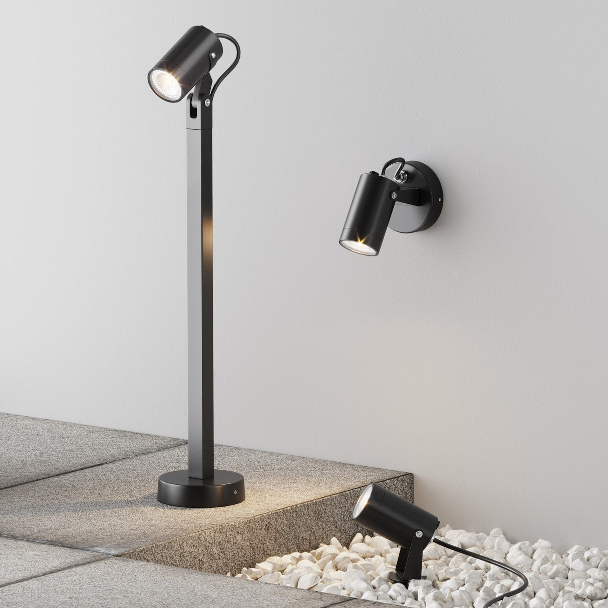 A modern garden is illuminated with a Scope Bollard Light 60cm-Black as part of an outdoor lighting setup, featuring three spotlights: one on a vertical pole, another wall-mounted, and the third on the ground highlighting white stones. The scene is minimal and contemporary.