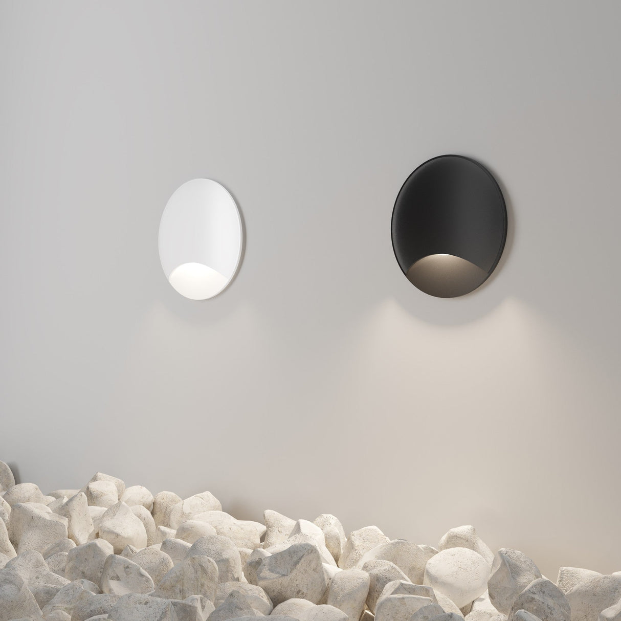 A minimalist wall showcases two Biscotti Outdoor 1 Beam LED Recessed Wall Lights—one white, one black—emitting soft glows. Smooth white stones below add texture. Ideal for outdoor lighting, its sleek design and IP65 rating ensure durability.
