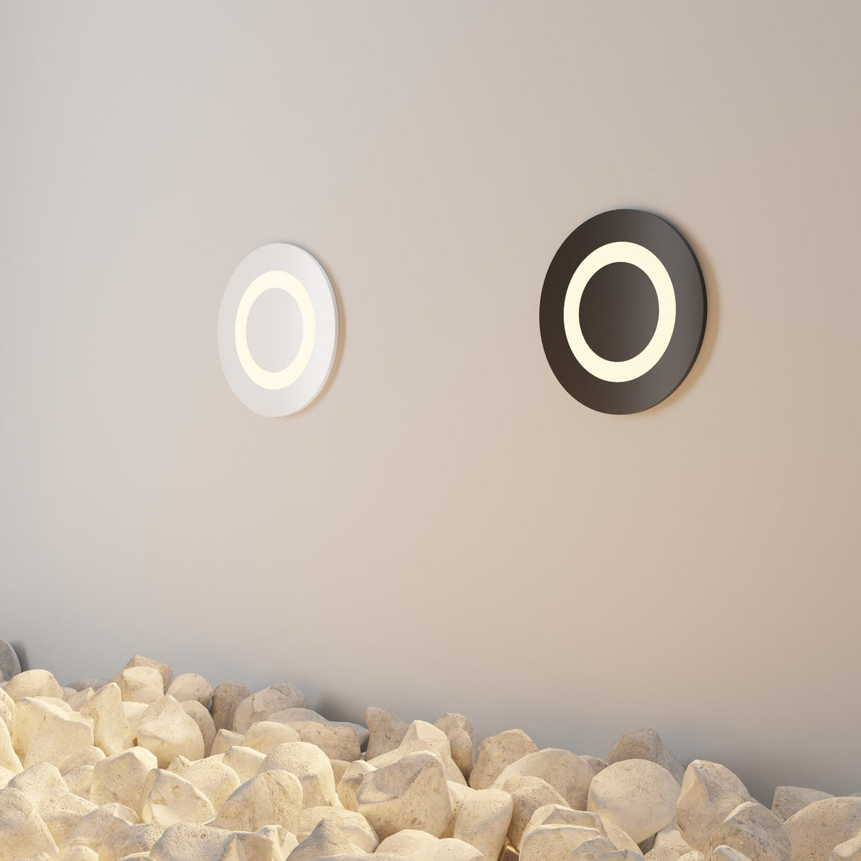 A minimalist wall displays two circular recessed lights, each casting a soft glow: one with a black ring, the other is the Limo Outdoor LED Recessed Wall Light - White. The floor features white decorative rocks that enhance the elegant settings texture.