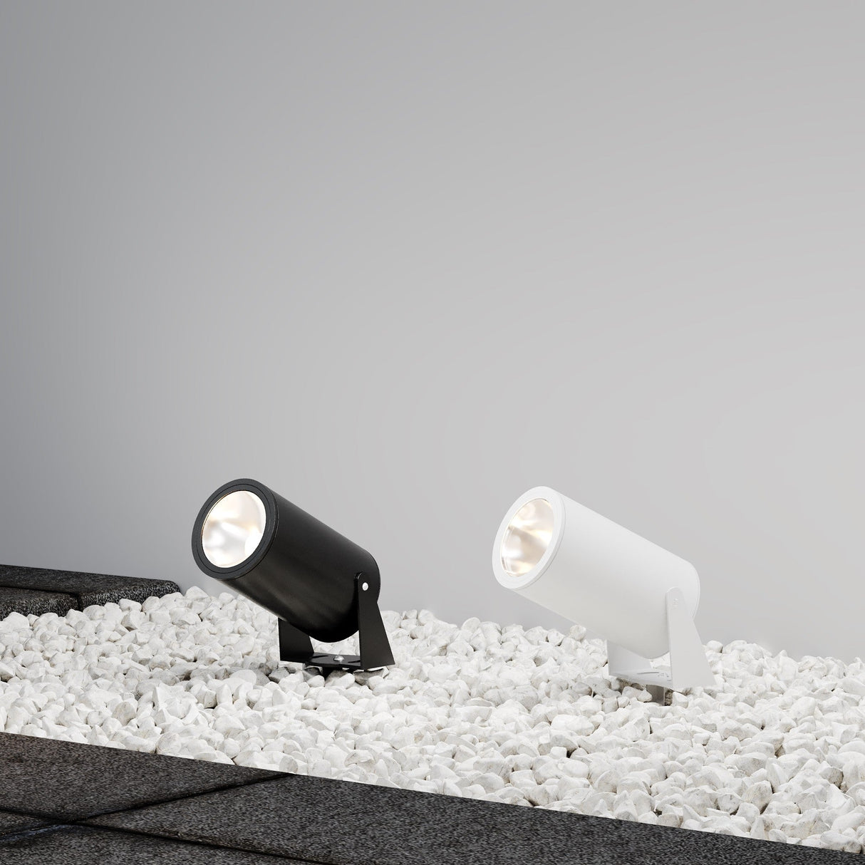 A Bern Outdoor LED Spike Light 30W 3000K in white accompanies a black spotlight, both featuring LED sources. Positioned on white pebbles, they cast a soft upward glow against a plain gray background.