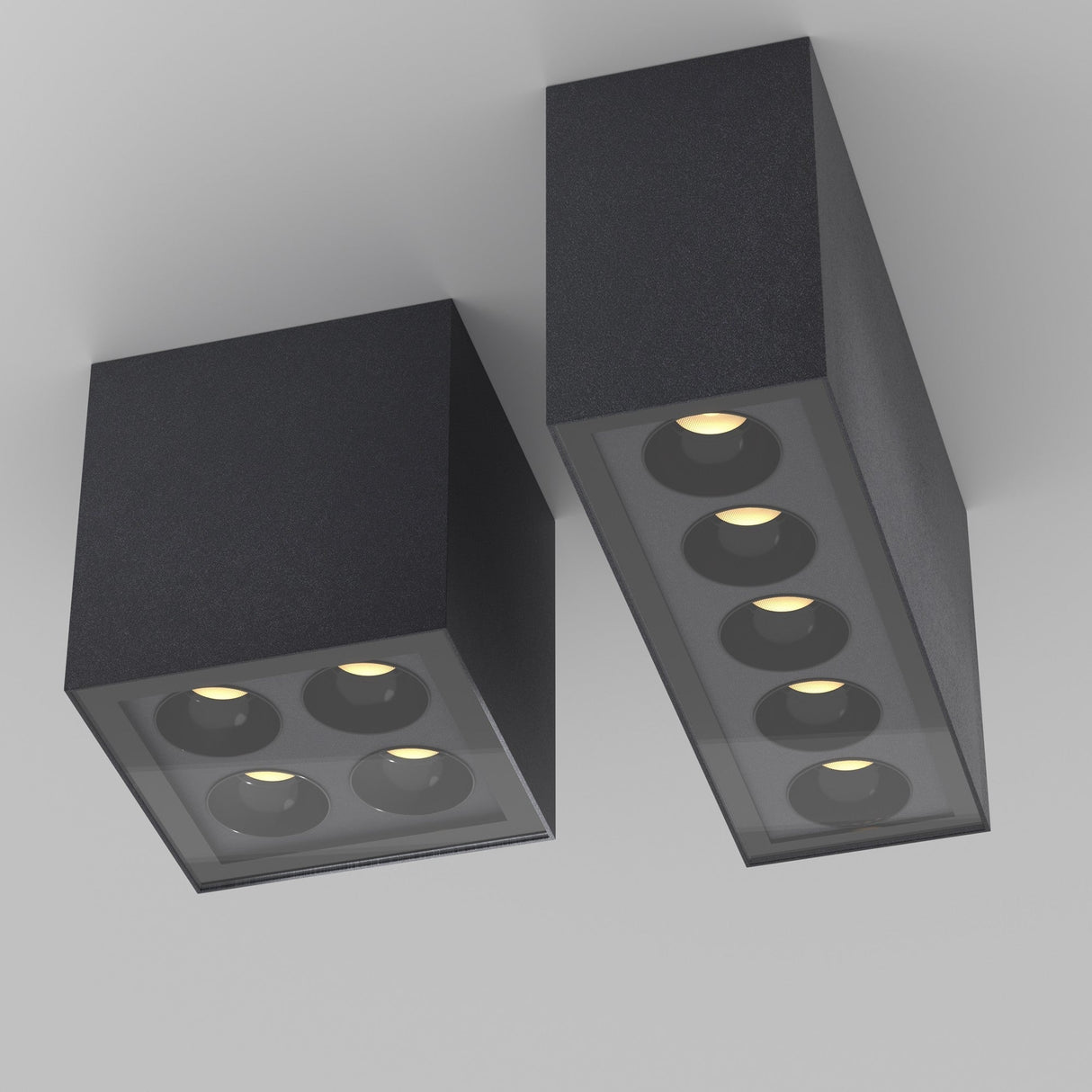 Two Ares Outdoor LED Square Downlights in graphite are installed side by side. Each minimalistic black fixture amidst the light-colored ceiling features multiple round bulbs that emit a subtle warm glow, ensuring energy-efficient lighting with a sleek look.