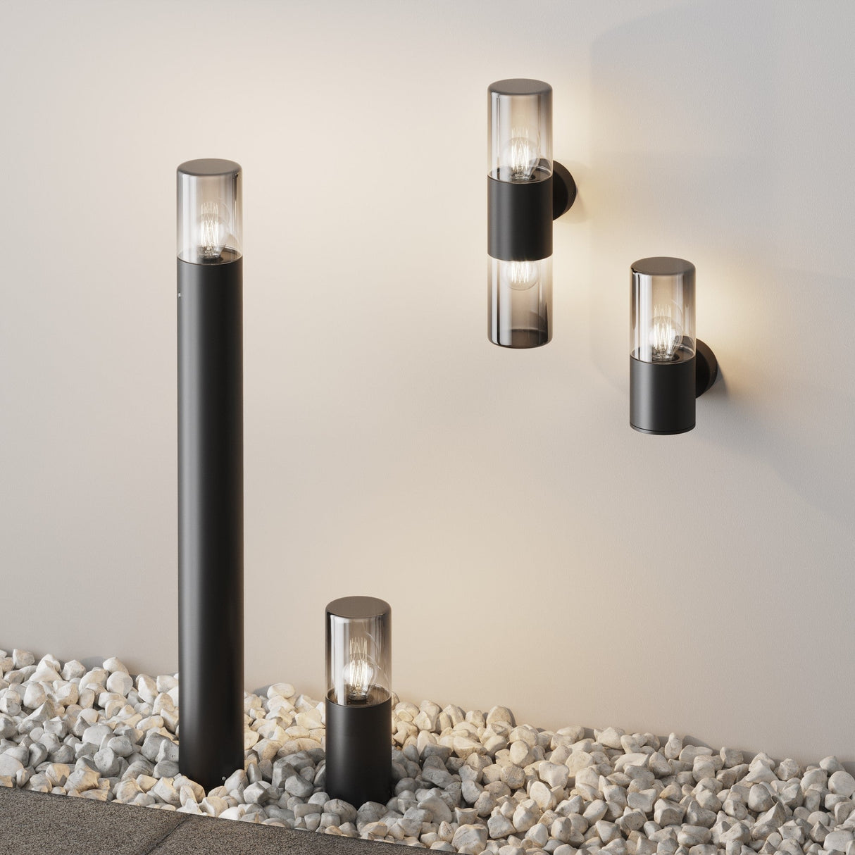 Outdoor lighting fixtures in a sleek graphite finish elegantly stand on a light-colored wall surrounded by small white stones. The setup features the Amas Bollard Light 80cm - Graphite, with tall and short post lights, plus two wall-mounted lights, all showcasing cylindrical glass covers and glowing bulbs.