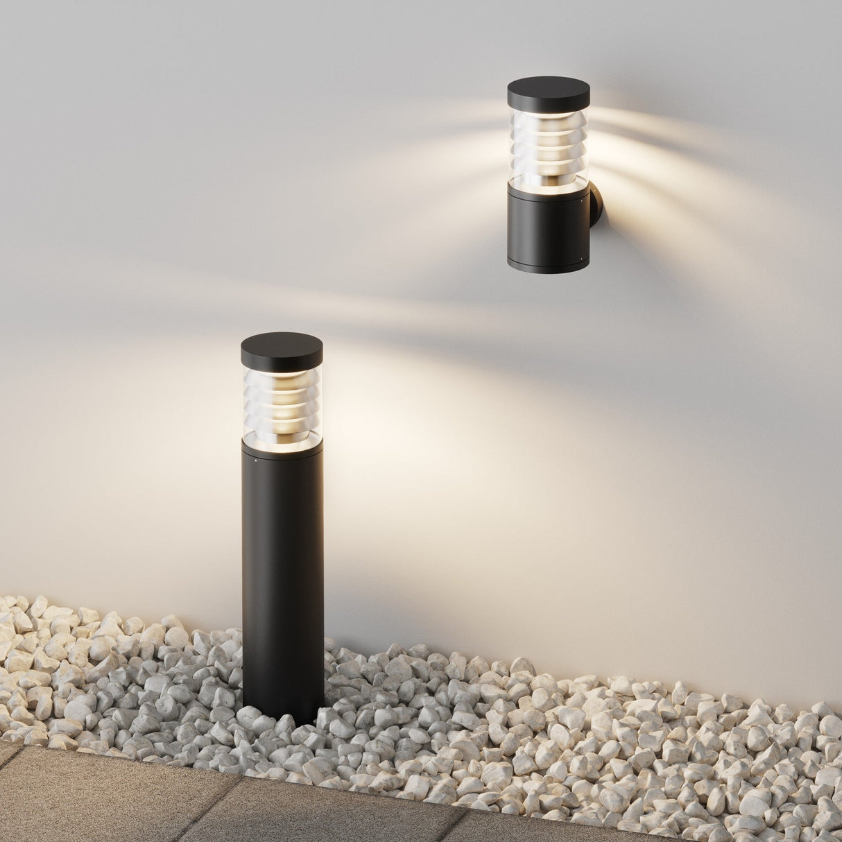 A black, cylindrical wall light and the Spir Bollard Light 3000K 12W - Graphite illuminate a gray surface. The post light stands on white pebbles beside paving stones, while the wall light casts a soft glow on the wall, enhancing the outdoor ambiance.