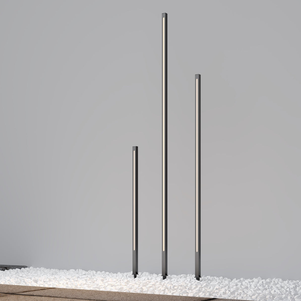 A trio of Pole LED Outdoor Spike Lights at 120cm in graphite stand on white pebbles against a plain gray backdrop, exuding sleek outdoor lighting elegance.