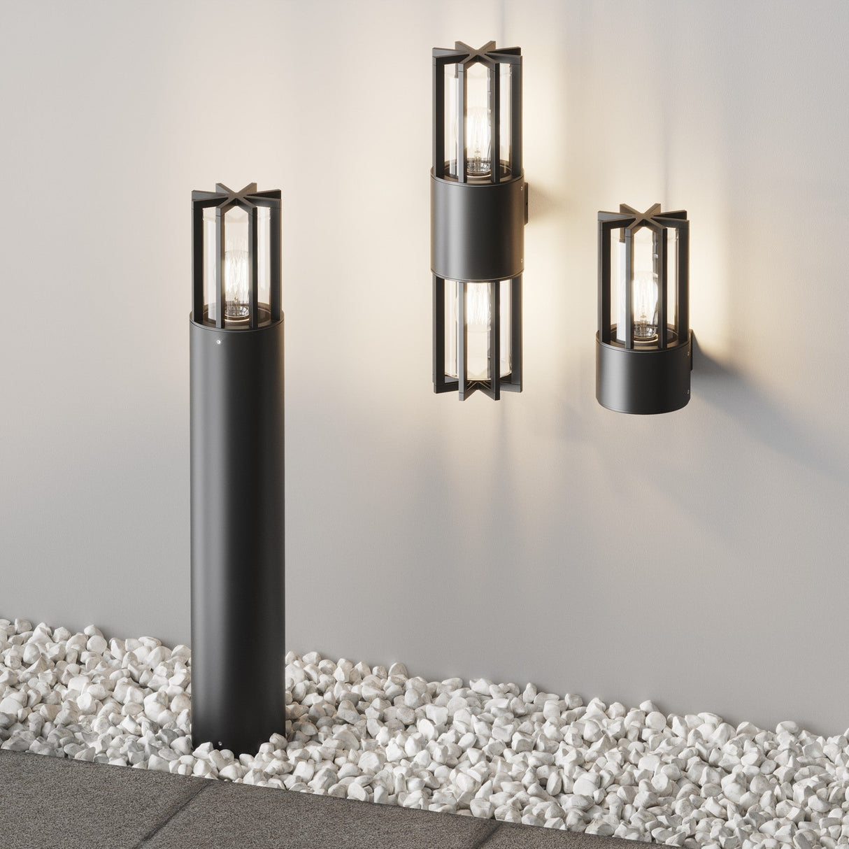Modern outdoor lighting with two lantern-style wall sconces and the Barrel Bollard Light 75cm in graphite on a textured wall, complemented by white decorative stones and paving stones in the foreground.