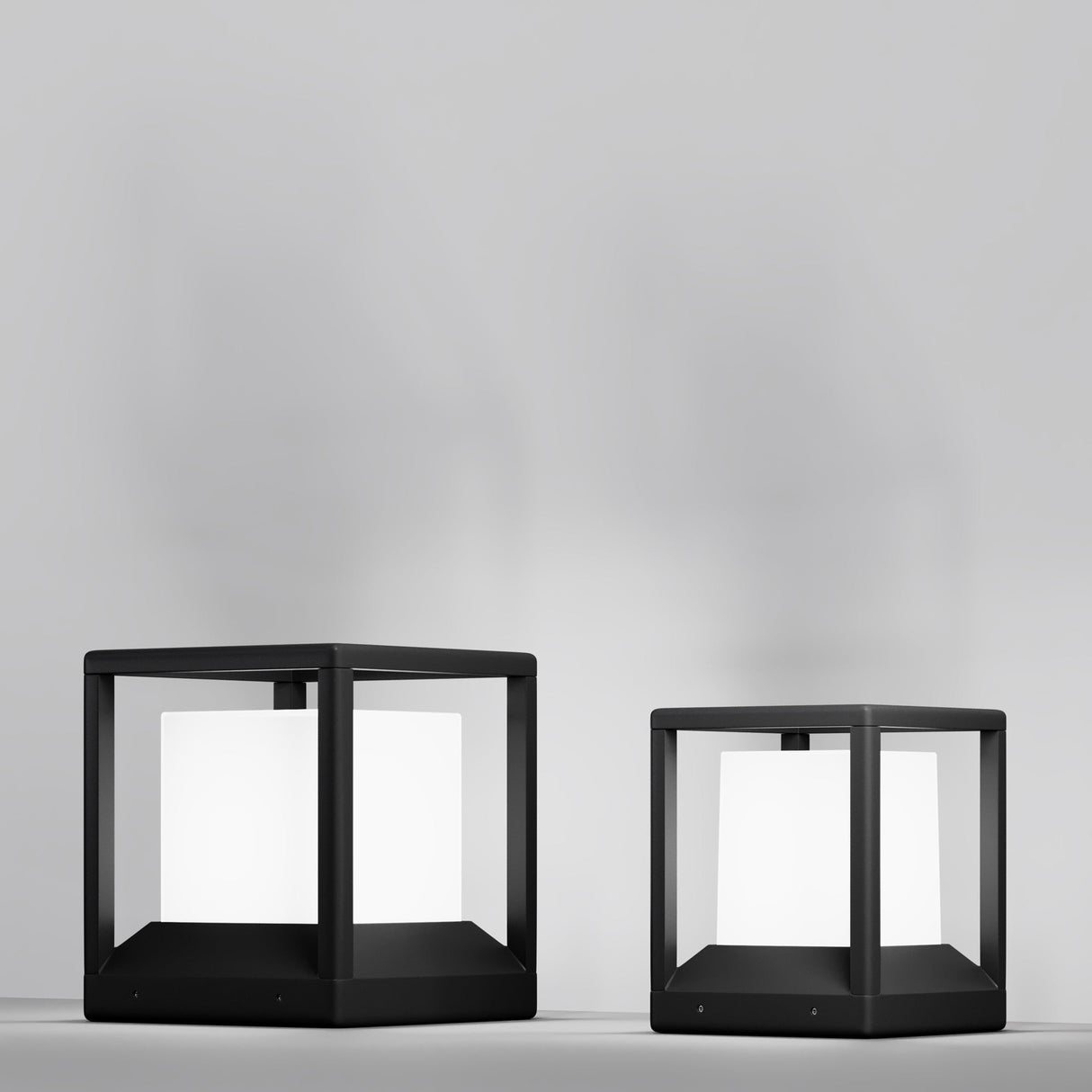 Two modern Trim Pedestal Lights with black frames and a sleek graphite finish sit on a smooth surface against a light gray backdrop. The taller light on the left complements the shorter one on the right, both offering an elegant, soft white glow for outdoor settings.