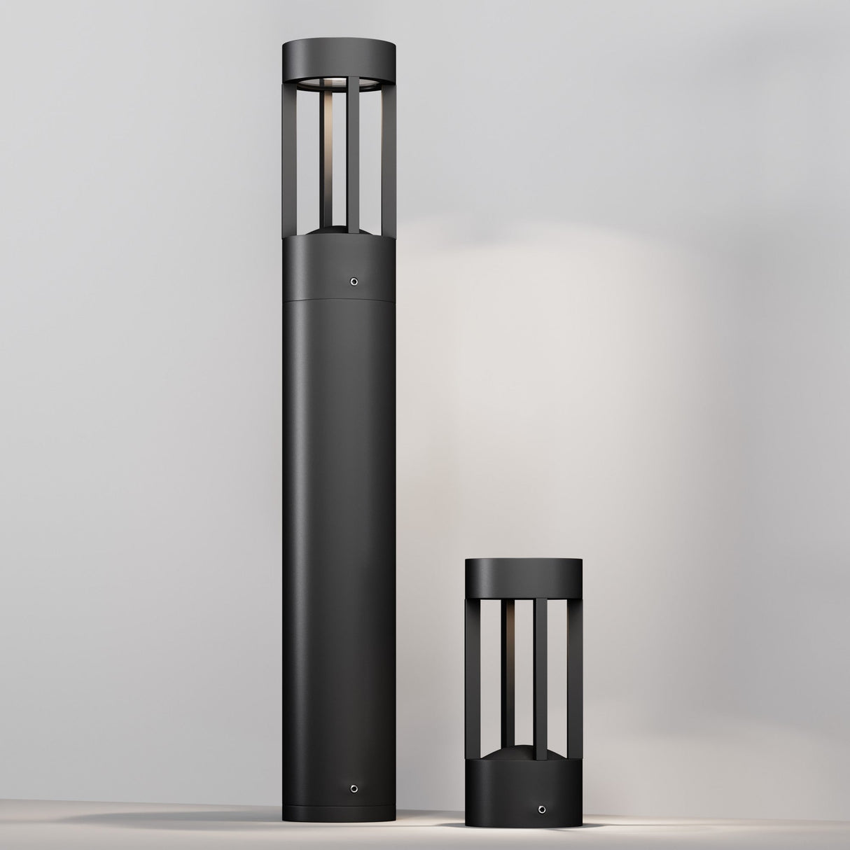 Two Vint LED Bollard Lights with minimalist graphite finishes stand on a light surface against a neutral background. The taller one on the left emits a soft glow, while the shorter one is on the right, both boasting sleek, cylindrical designs for a sophisticated look.