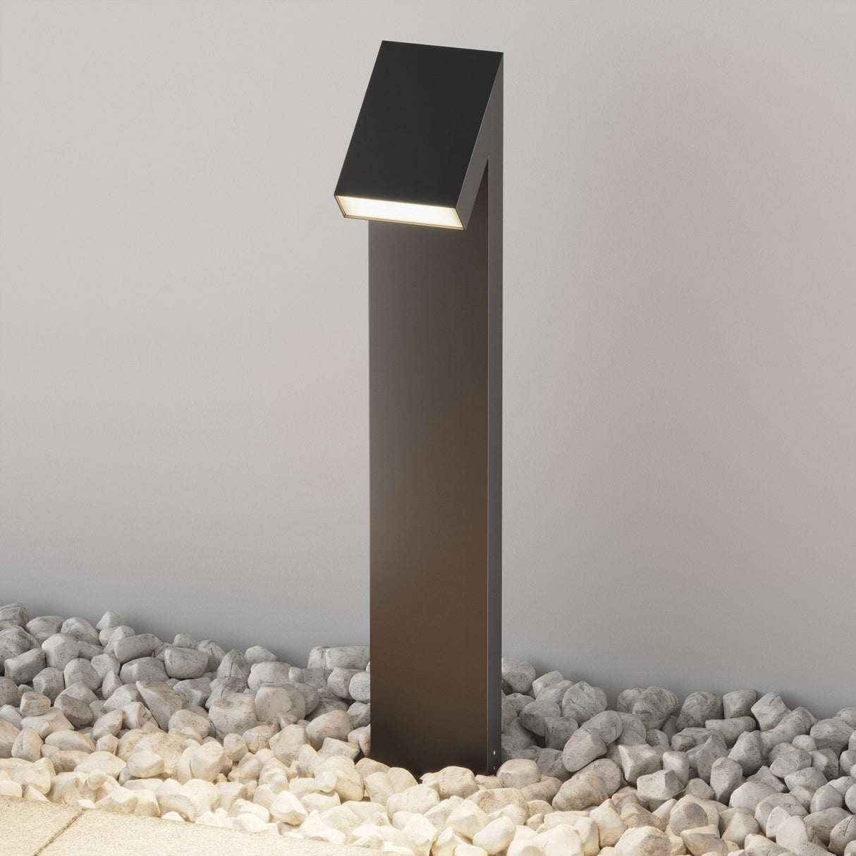 A contemporary Guss LED Bollard Light 90cm in graphite stands on a gravel surface, emitting a warm LED glow downward. IP65 rated for durability, it is against a plain beige wall, surrounded by small gray and white stones.