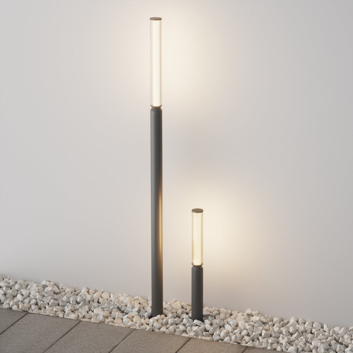 Two modern lights with a sleek graphite finish and glass tops illuminate against a white wall. Surrounded by decorative stones near pavement, the taller Lit LED Short Bollard Light - Graphite subtly stands behind the shorter one, casting a soft glow.
