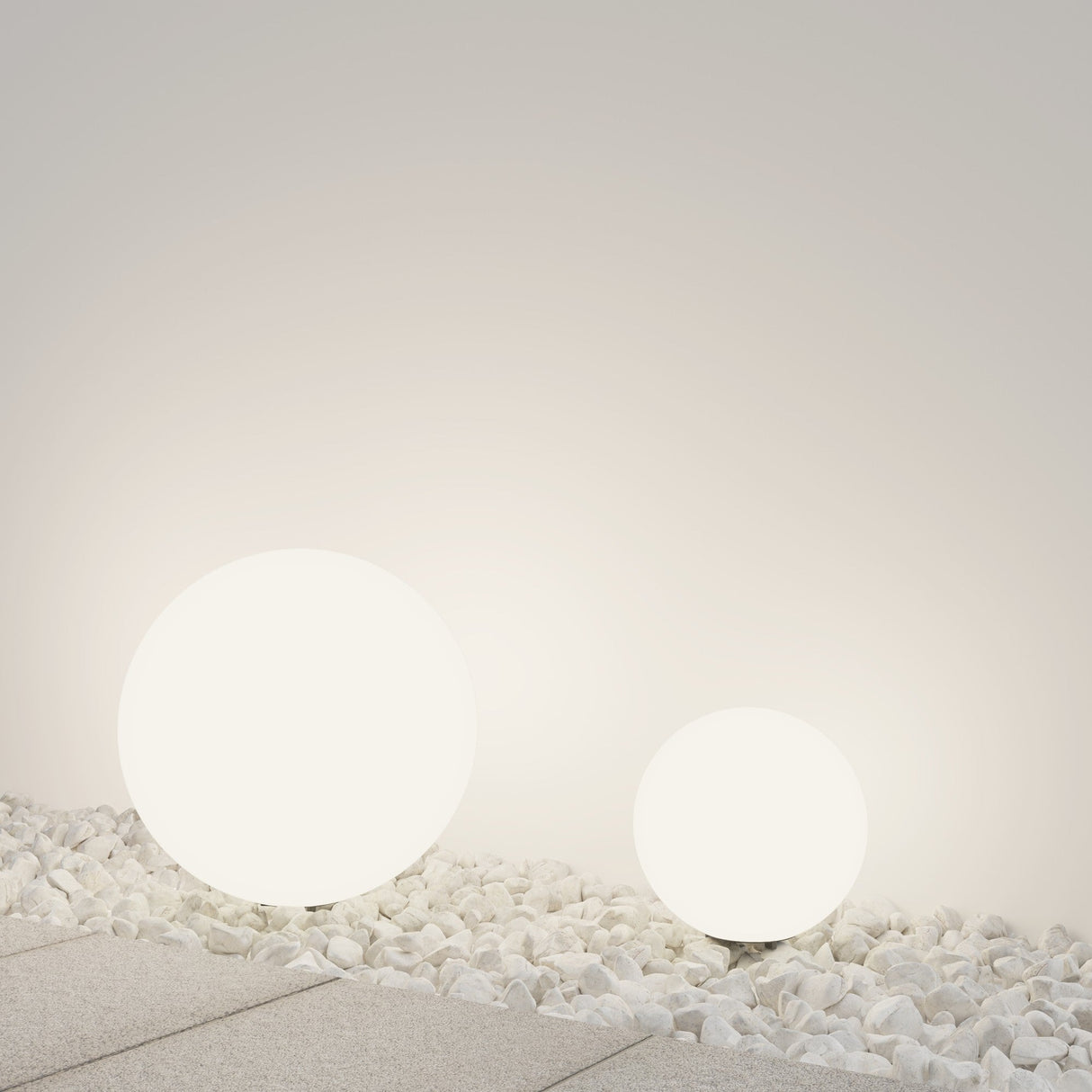 The Erda Pedestal Light 29cm - White and a larger spherical lamp illuminate white pebbles on a smooth surface, casting a soft glow against the light gray wall. This weather-resistant outdoor lighting beautifully enhances any serene setting.