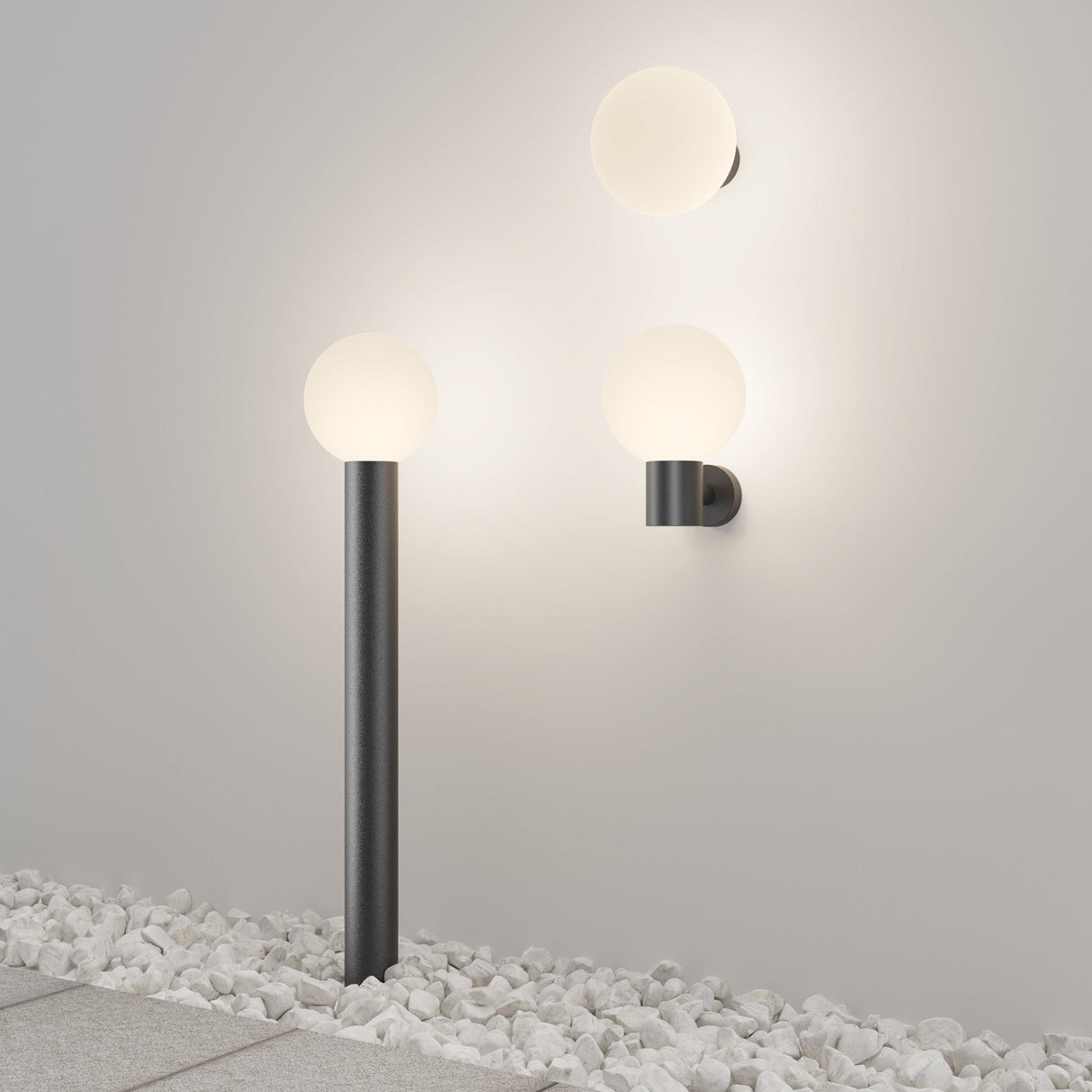 The lighting design includes two Bold Outdoor Wall Lights - Black (O598WL-01B1) with round bulbs and a matching floor lamp, all on sleek black stands. The IP65-rated lights are set against a light gray wall with white pebbles below, offering durability and style in diverse conditions.