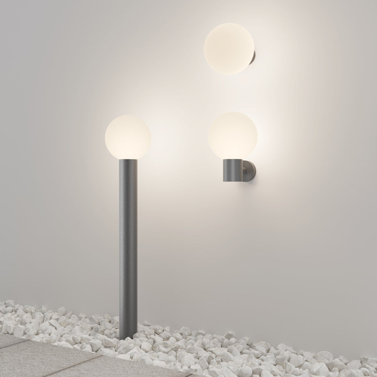 The Bold Outdoor Wall Light in Grey (O598WL-01GR1) features spherical frosted glass globes mounted on a wall and placed on a stone-covered ground. Its weather-resistant metal construction and minimalist design cast a soft glow on the white wall.