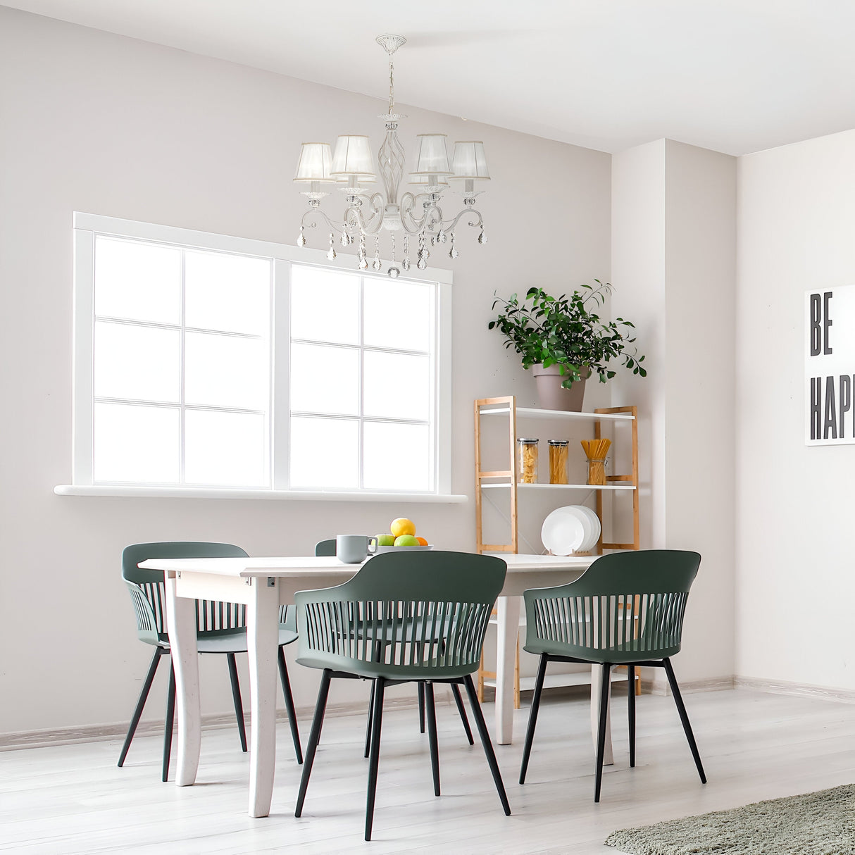 In a bright dining space, a white table is accompanied by four dark green chairs. The Grace 6 Light Chandelier in white casts elegant light above. A fruit bowl with lemons and apples rests on the table. Nearby, a wooden shelf displays plants and dishes, while a BE HAPPY sign adorns the wall.