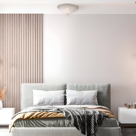 A modern bedroom includes a gray upholstered bed, two plaid pillows, and a plaid blanket. Light wooden paneling adorns the left wall. The ceiling is graced by the Pascal 3 Classic Light Flush in white and gold, with a small table and decor on the right.