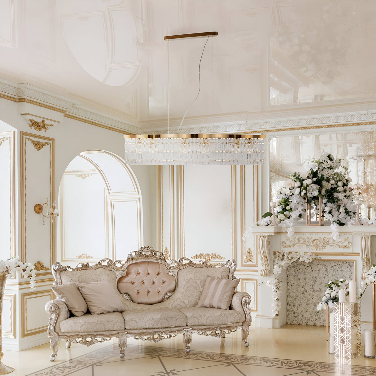 Luxurious living room with ornate white and gold decor, featuring an elegant couch with plush cushions. The Dune 6 Light Island Suspension - Gold hangs above, adorned with crystal accents. A floral arrangement decorates the white mantle in this opulent, classical interior.