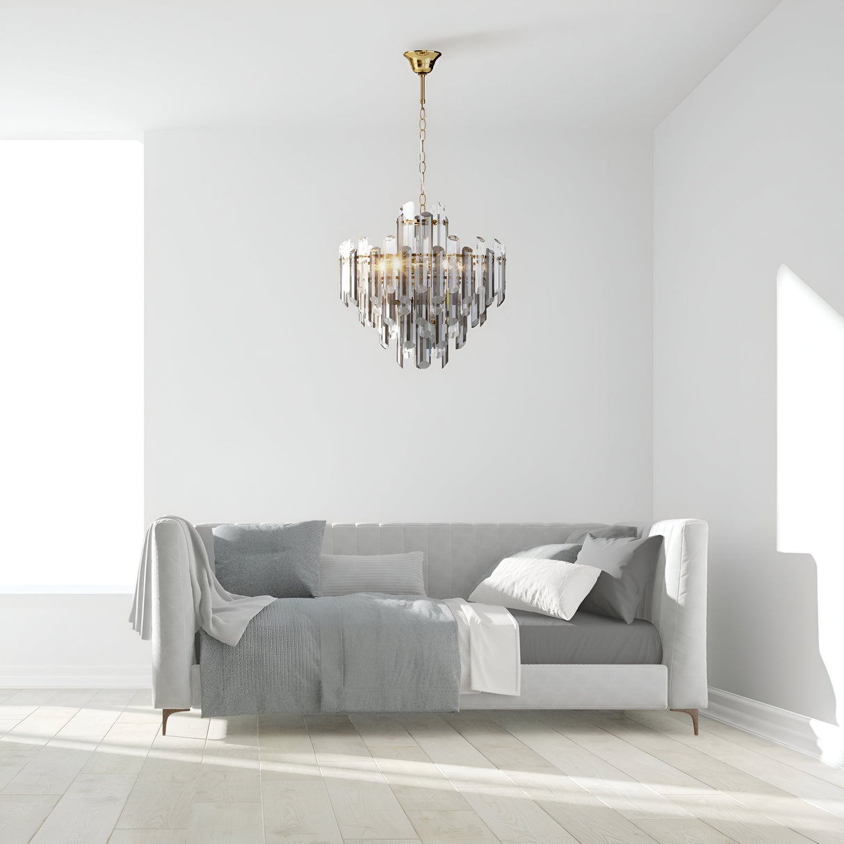 A minimalist white room has a plush white sofa adorned with gray and white pillows and a draped blanket. A Flare 6 Light Chandelier - Gold with faceted crystals hangs from the ceiling, shimmering in the natural light filtering through the window, illuminating the space.