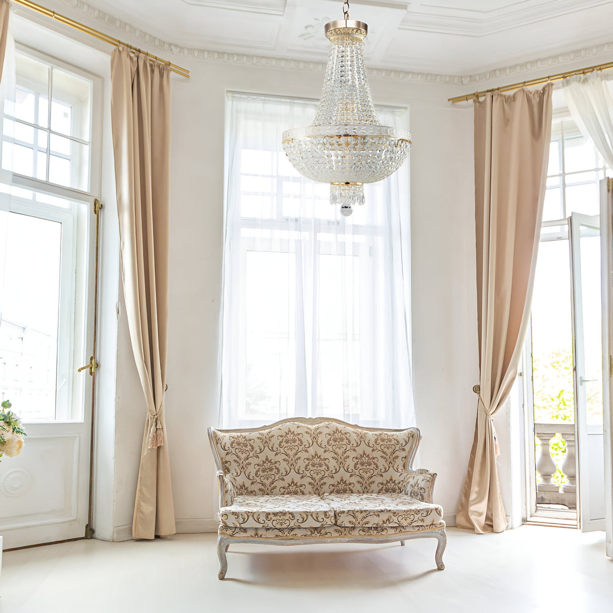 An elegant room is bright and airy with large windows, sheer white curtains, and beige drapes. A vintage sofa with ornate upholstery sits beneath the Bella 6 Light Crystal Chandelier - Gold, exuding classic sophistication through its luxury lighting.