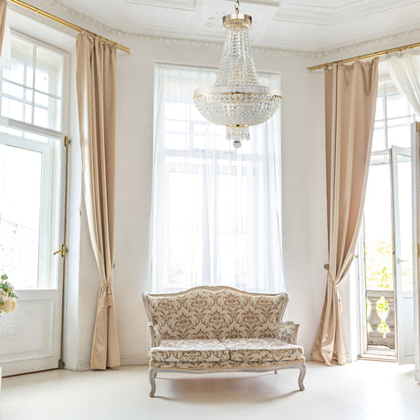 An elegant room is bright and airy with large windows, sheer white curtains, and beige drapes. A vintage sofa with ornate upholstery sits beneath the Bella 6 Light Crystal Chandelier - Gold, exuding classic sophistication through its luxury lighting.