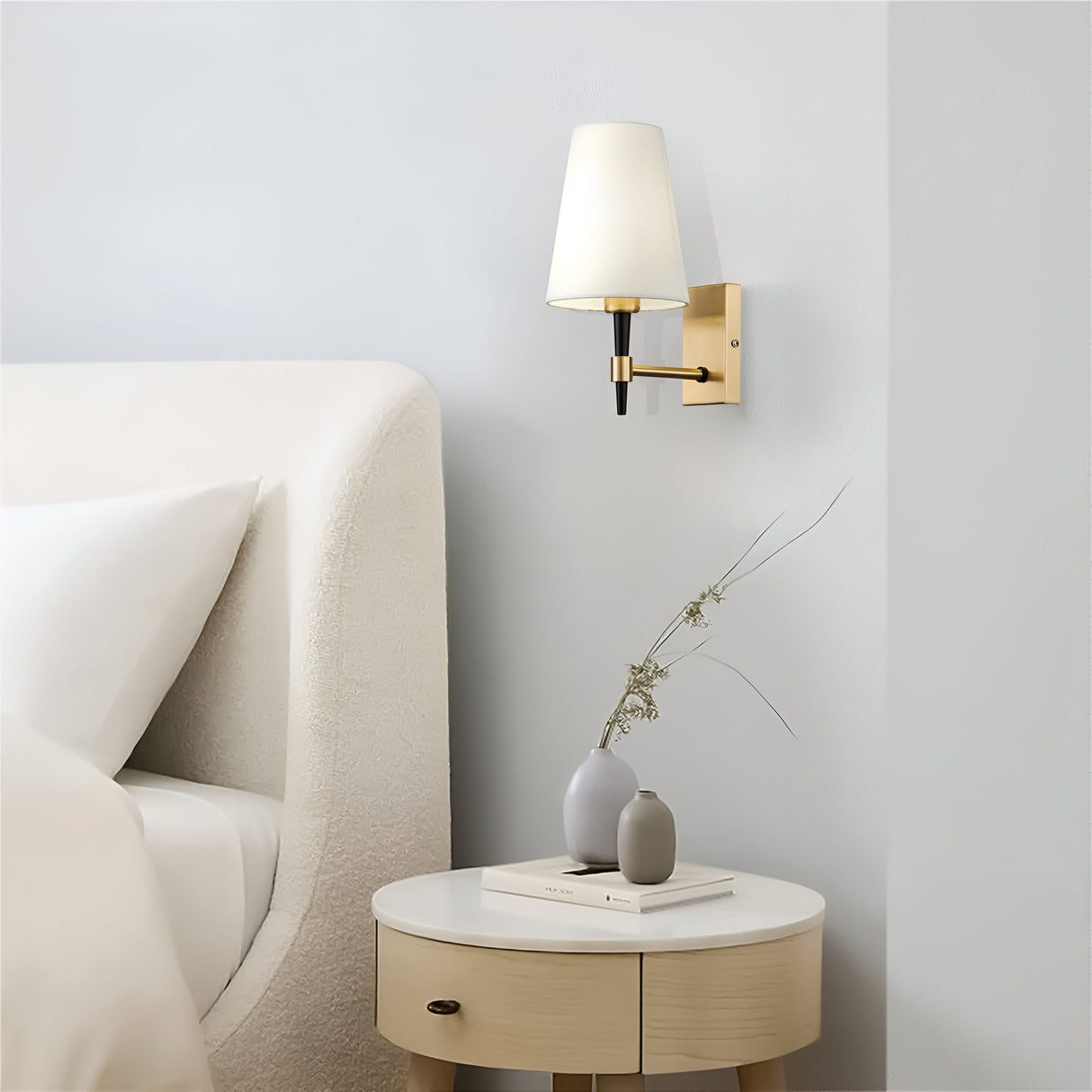 A minimalist bedroom includes a beige upholstered bed with white bedding. The Zaragoza Wall Light - Brass With White Fabric Shade adds ambiance above a small round bedside table, which holds a closed book and gray vase with dried flowers.