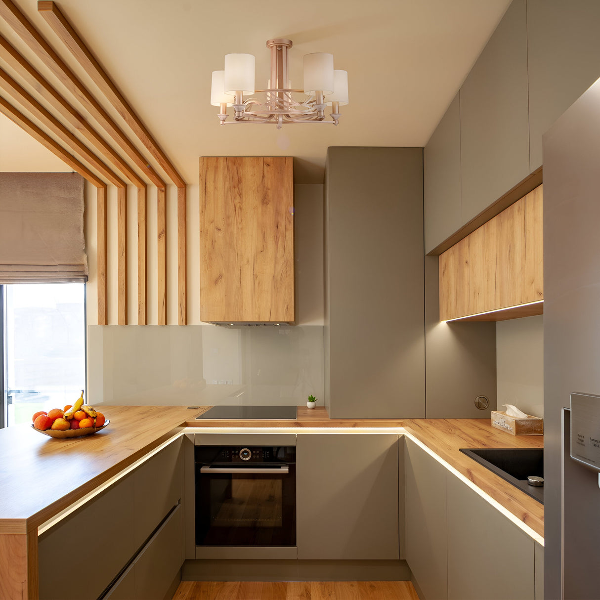 A modern kitchen features gray cabinets and wood accents. A built-in oven is beneath a wooden countertop, next to a small sink. A fruit bowl with bananas and apples sits on the counter, while a Vittoria 5 Light Chandelier - Gold elegantly hangs above.