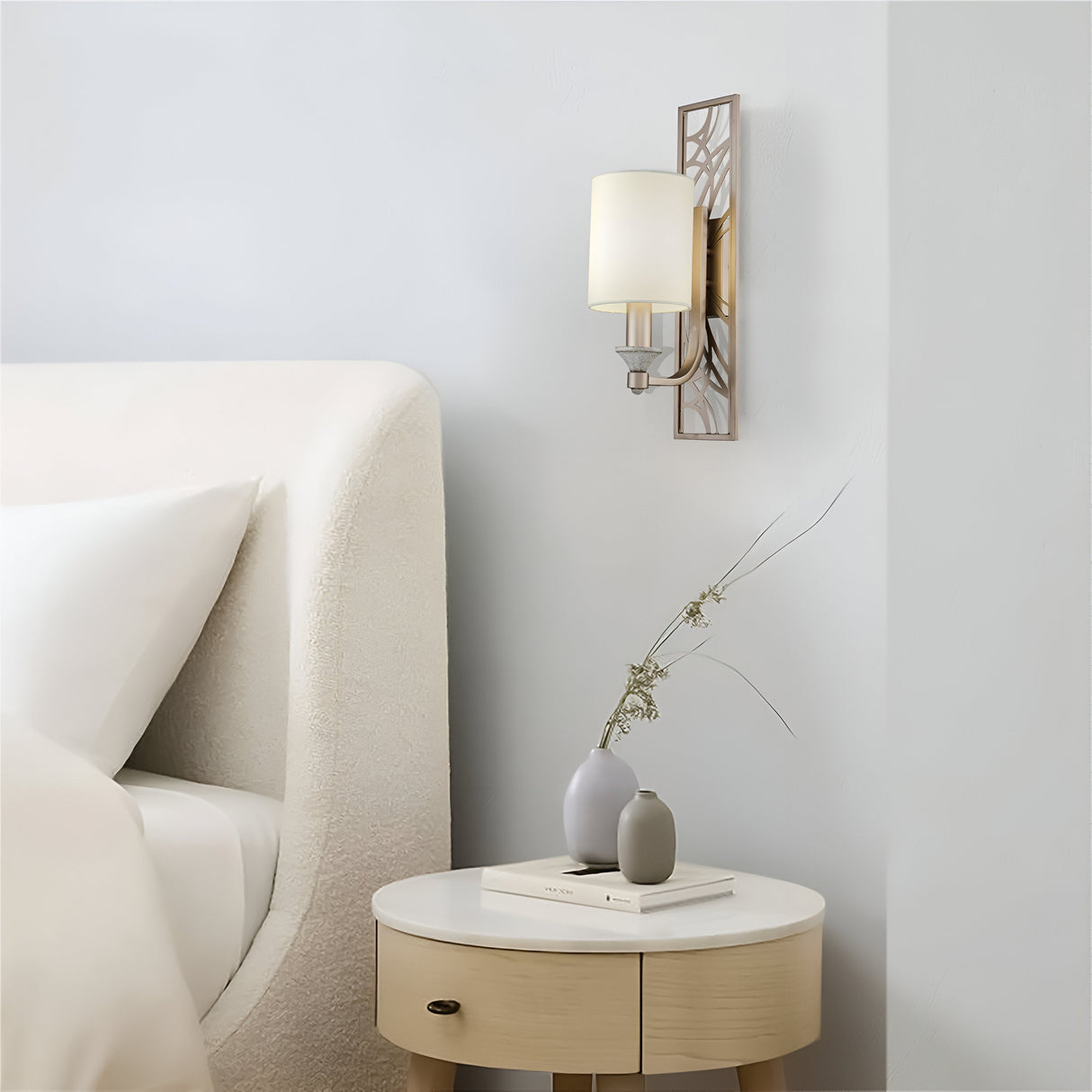 A modern bedroom showcases a white curved headboard and a small round bedside table with a gray vase of dried flowers and stacked books. The Vittoria Wall Light - Gold With Cream Fabric Shade softly casts ambient lighting on the neutral-toned wall.
