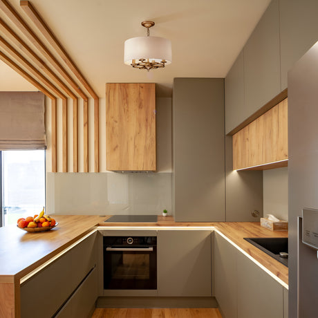 A modern kitchen features matte gray cabinets, wooden countertops, and a built-in oven. A fruit bowl with bananas and apples rests on the counter. The Anna 4 Light Chandelier - Gold casts an ambient glow from above, while vertical wooden slats adorn one wall.