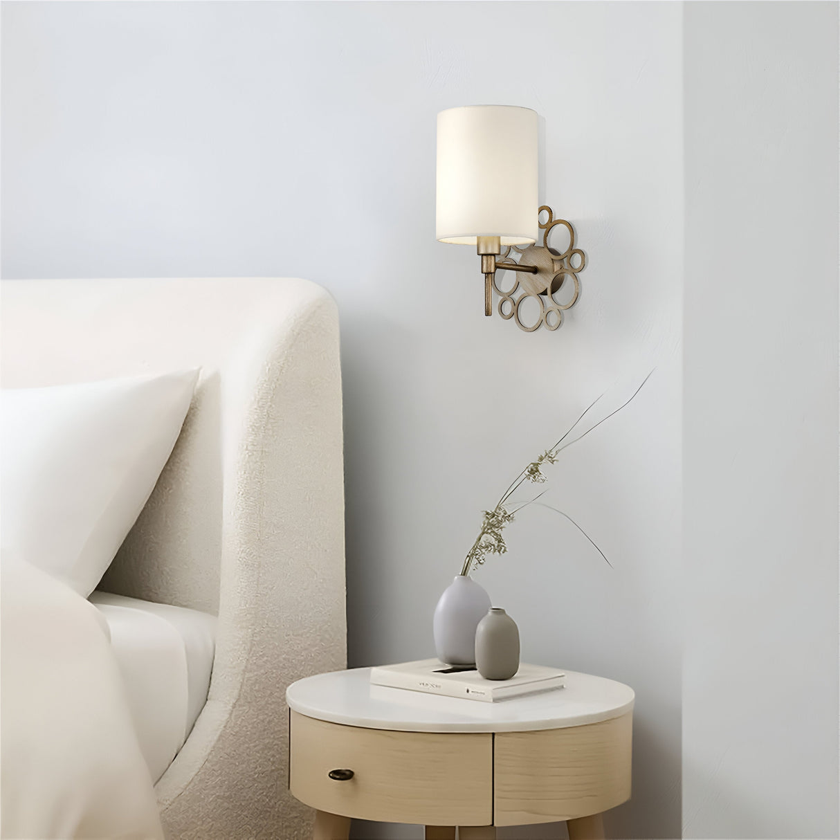 A modern bedroom features a curved upholstered bed, a round nightstand with a gray vase of dried flowers and two books, and the Anna Wall Light in Antique Gold with Cream Shade mounted on a light-colored wall.