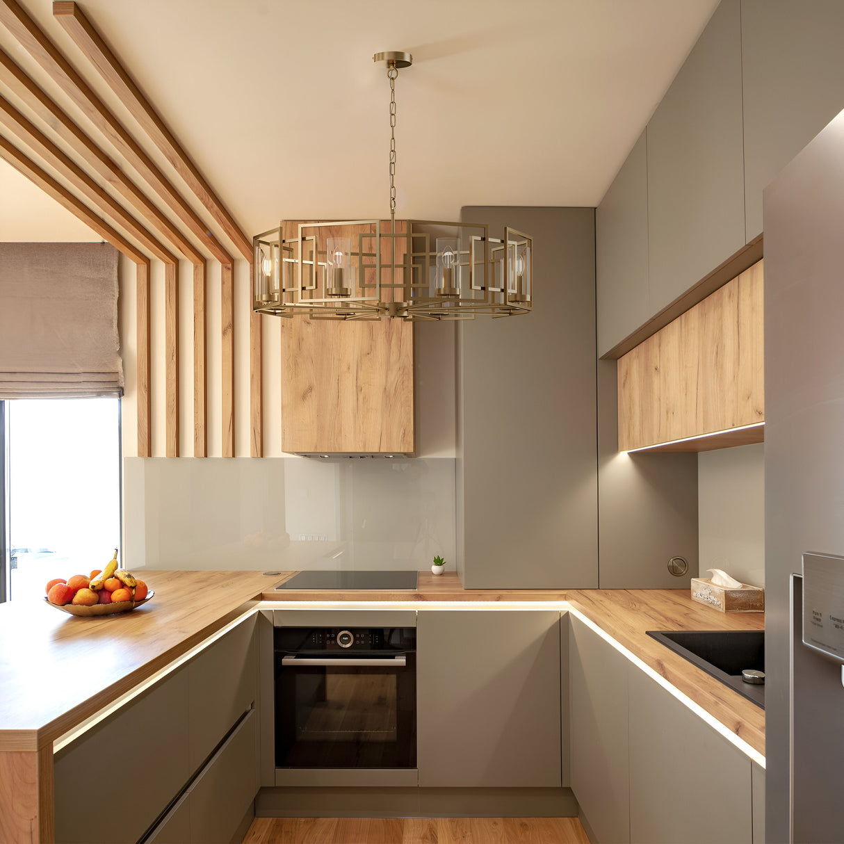 Modern kitchen featuring L-shaped wooden countertops, light gray cabinets, and a black built-in oven. The Bowi 8 Light Chandelier - Gold adds elegance overhead. A fruit bowl with bananas and apples rests on the counter near a window with vertical wooden accents enhancing the natural lighting.
