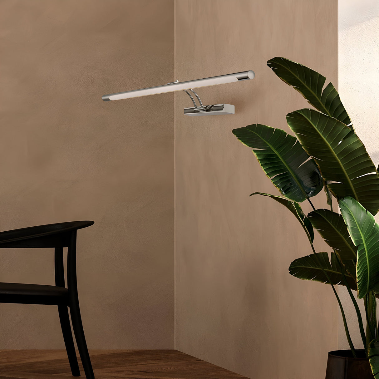 A modern interior showcases the Fino LED Picture Light 12.1W 3000K - Chrome, casting light on a beige textured wall. Close by, a large green potted plant sits on the wooden floor next to a partially visible black chair with a curved back.