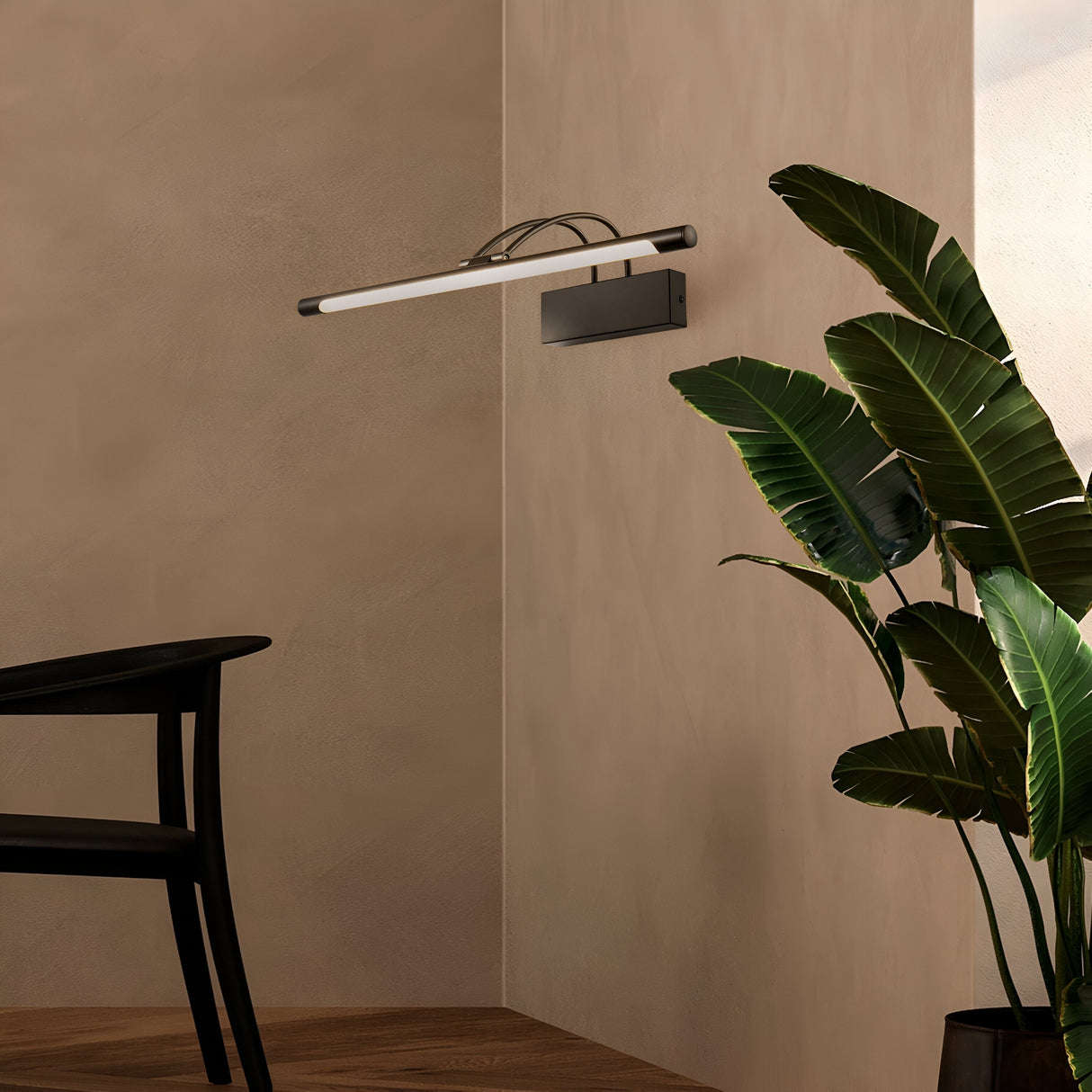 A minimalistic corner features the sleek Finelli LED Picture Light 12.1W 3000K - Black & White, elegantly mounted above a large potted plant with broad green leaves. In the foreground, a black chair with wooden accents is partially visible against soft beige walls framing this serene setting.