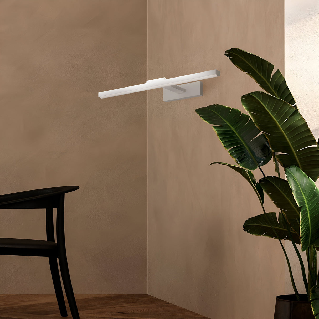 A minimalist room corner features a Bris LED Picture Light 12W 4000K - White, complementing the white wall-mounted fixture. Nearby, a large green plant stands on a wooden floor with part of a black chair visible against the beige wall, enhancing the calm, modern ambiance.
