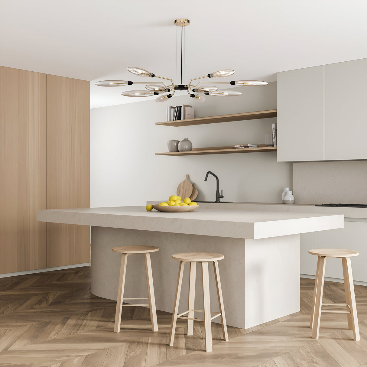 A modern kitchen with a minimalist design features a spacious island with three wooden stools. The Ventura 12 Light Chandelier - Gold adds elegance overhead. Wooden floors and shelves contrast with white cabinetry, while a bowl of lemons offers a vibrant pop of color.