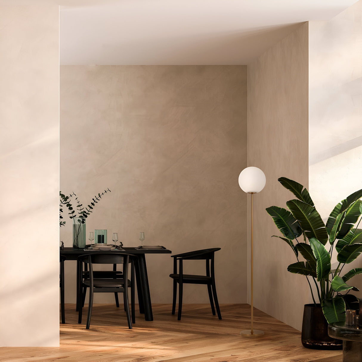 Minimalist dining area featuring a rectangular black table, two black chairs, and the Ring Floor Lamp in brass with a frosted glass globe. Potted plants bring life to the space, as beige walls and wooden flooring enhance the serene atmosphere and modern lighting style.