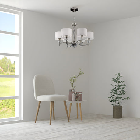 A minimalist room features a cushioned chair and a round table with potted plants. The Alicante 6 Light Chandelier - Nickel adds elegance as sunlight floods through the large window, framing lush greenery and epitomizing modern interiors.