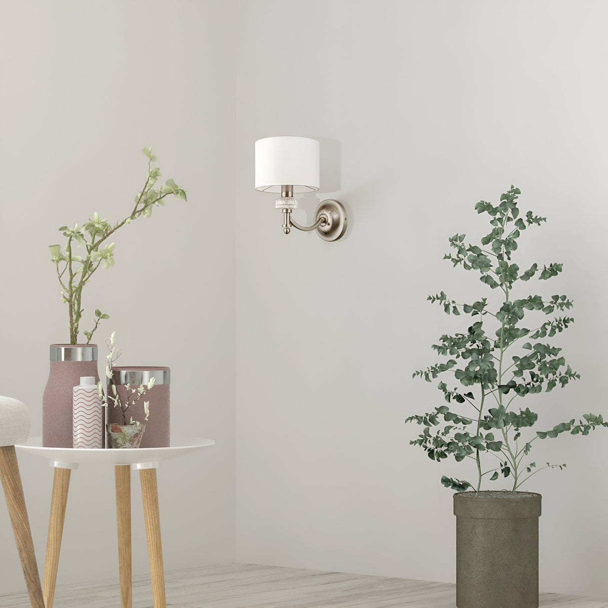 A minimalist corner showcases an Alicante Wall Light - Nickel with White Fabric Shade, a small round table with two pink vases containing branches, and a tall green plant in a gray pot on light wooden flooring, all beautifully lit by elegant lighting.