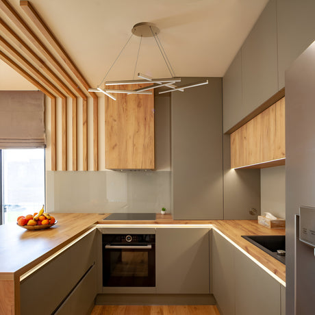 A modern kitchen features light wood countertops and cabinets, accented by the Line LED Suspension Light 4000K in Matte Gold. This geometric chandelier casts a warm glow above the center. Neutral tones prevail, with a fruit bowl on the counter and visible built-in oven and sink, plus vertical wooden accents on one wall.