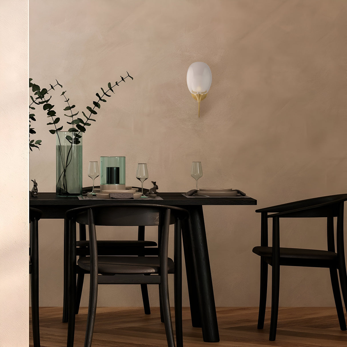 A modern dining area with a black table and two chairs showcases elegant place settings and glassware. A tall vase with eucalyptus branches adorns the table, while the Aura Wall Light - Brass, featuring a double-blown glass lampshade, casts ambient lighting. The space feels calm and inviting.