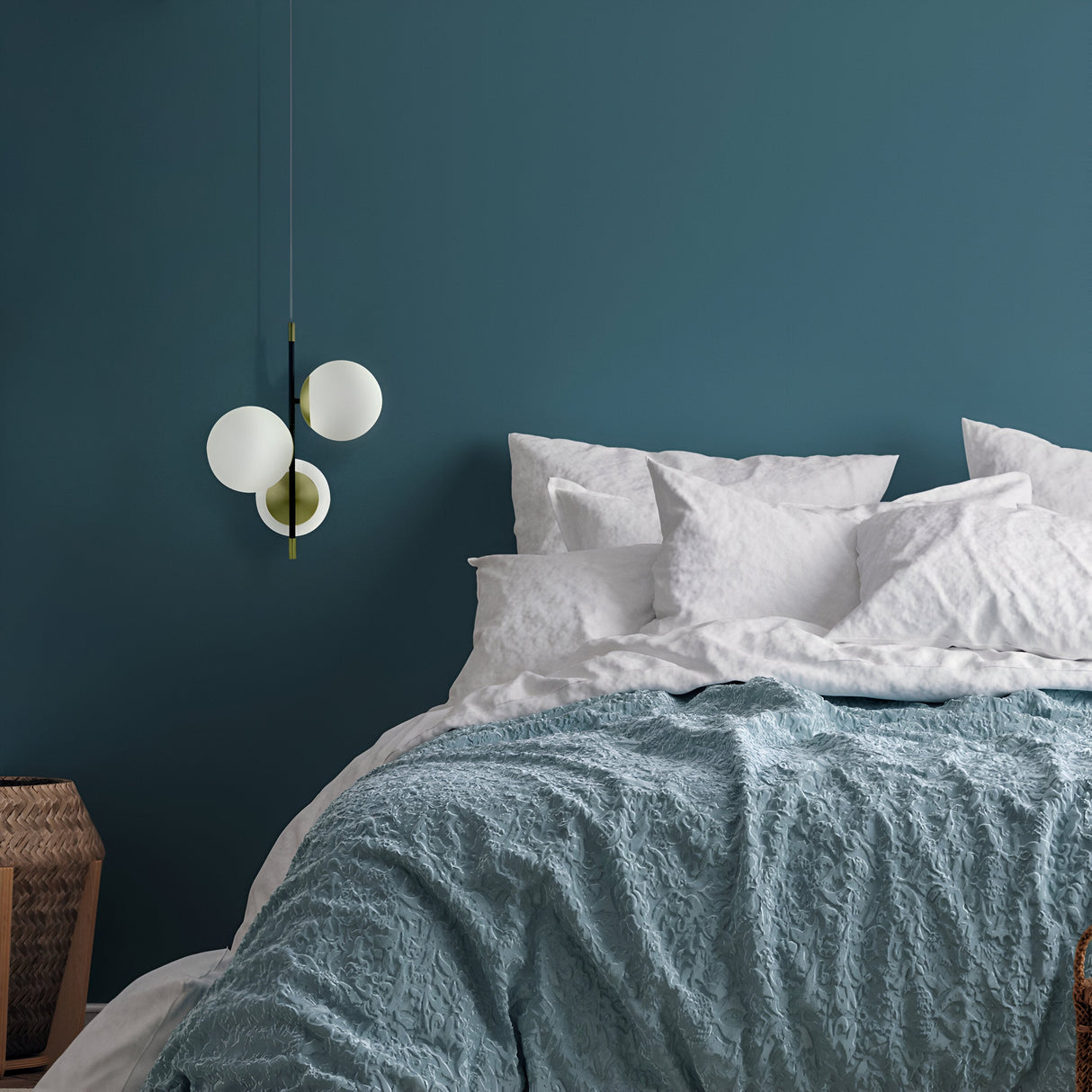 In a cozy bedroom, a bed with white pillows and a textured teal blanket sits against a teal wall. The Nostalgia 3 Light Pendant - Black & Gold With Frosted Glass casts a warm glow overhead. A wicker basket beside the bed enhances the rooms inviting charm.